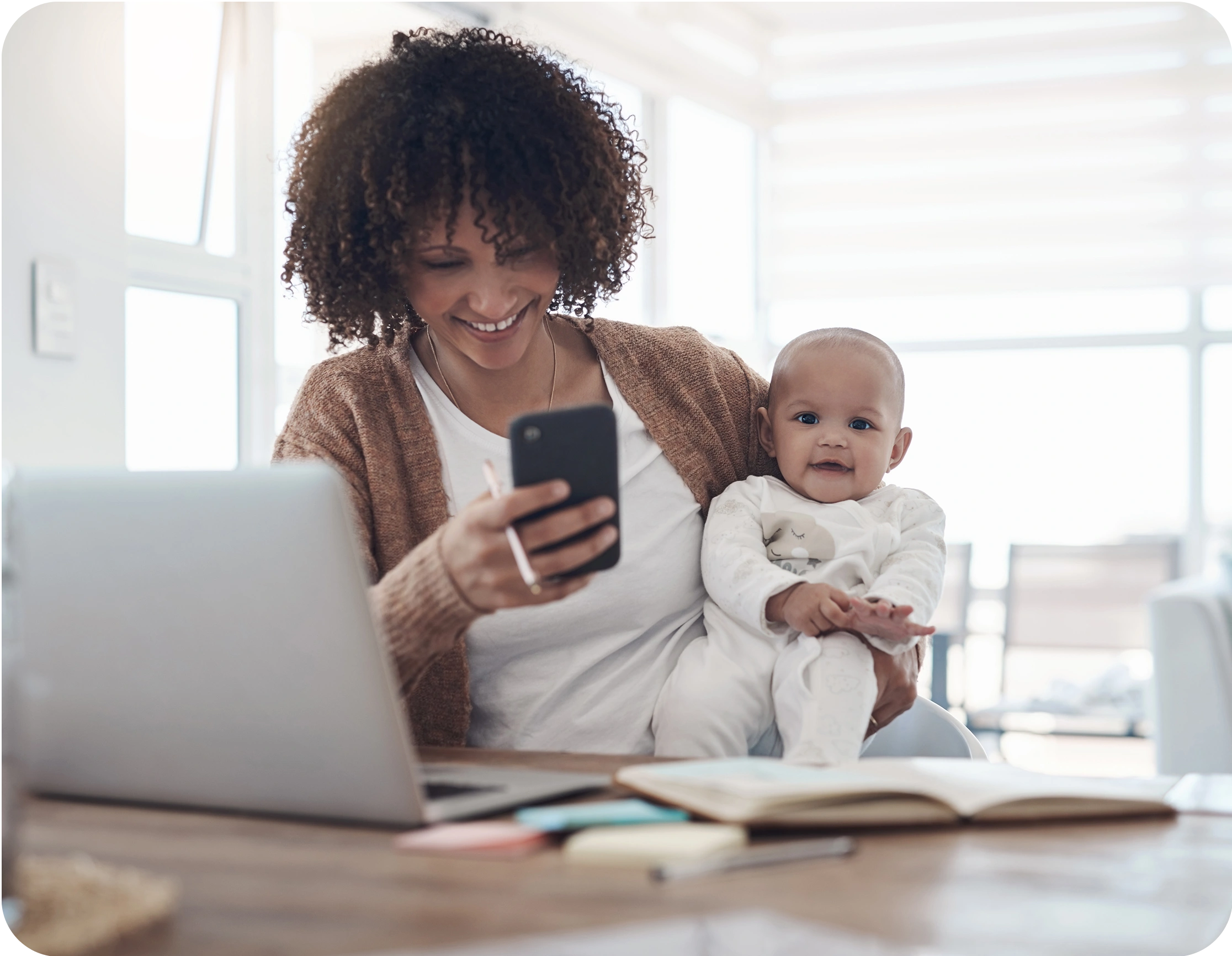 Mother holding a baby while using a smartphone, with a laptop and notebook on the table.