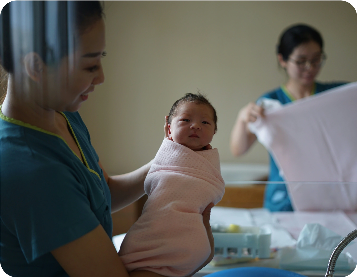 Healthcare worker holding a newborn wrapped in a pink blanket in a hospital setting.